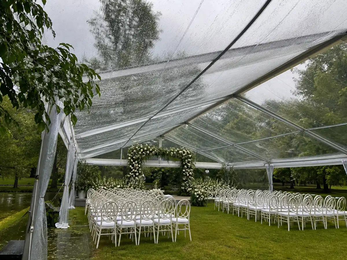 เต็นท์ใสกางคลุมโซนพิธีกลางสวนขณะฝนปรอย โดยยังมองเห็นต้นไม้และท้องฟ้าผ่านหลังคา