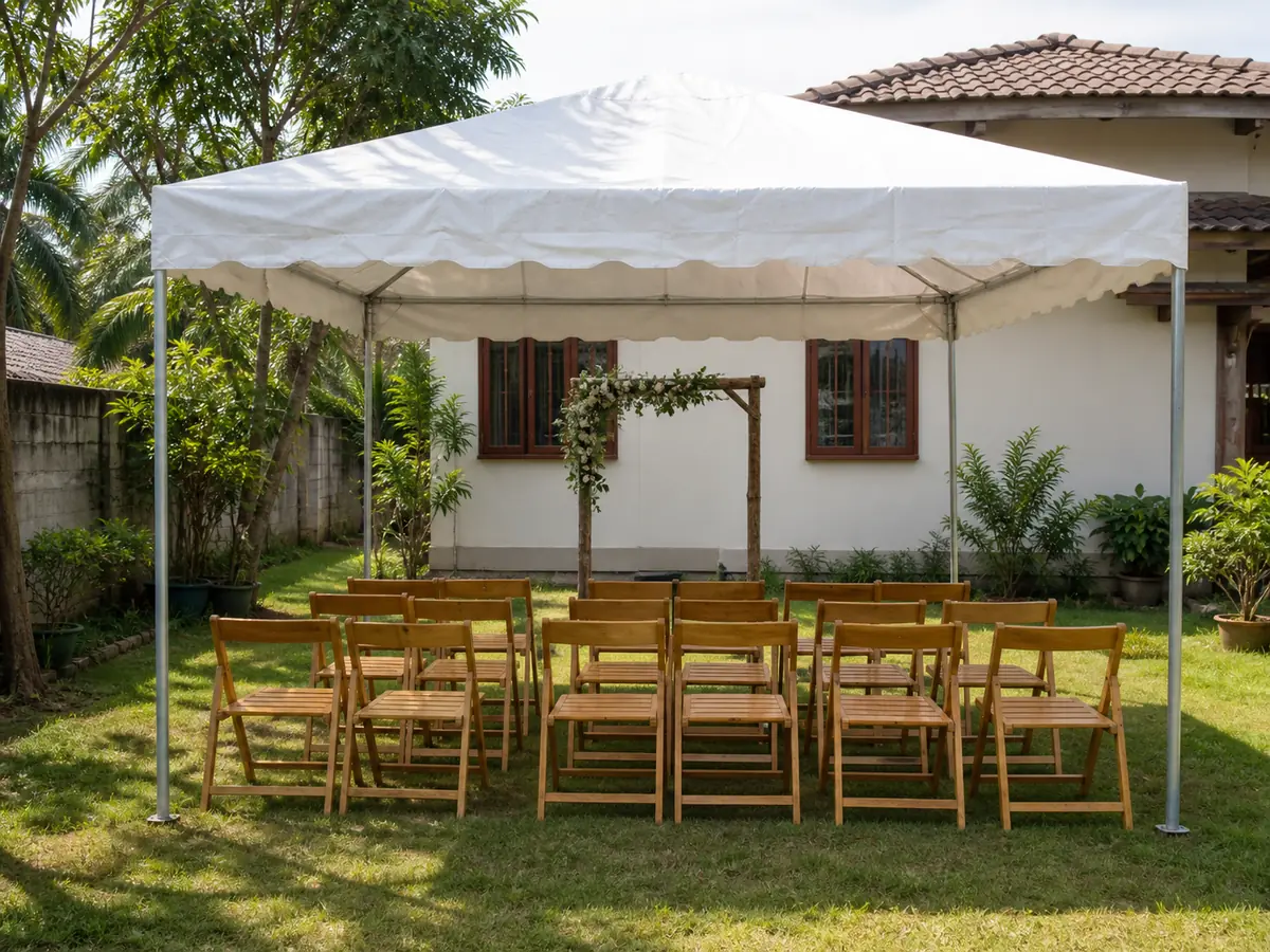 A home front lawn set up as a wedding venue with a rented white canopy, rows of plain wooden chairs and a simple greenery-dressed arch