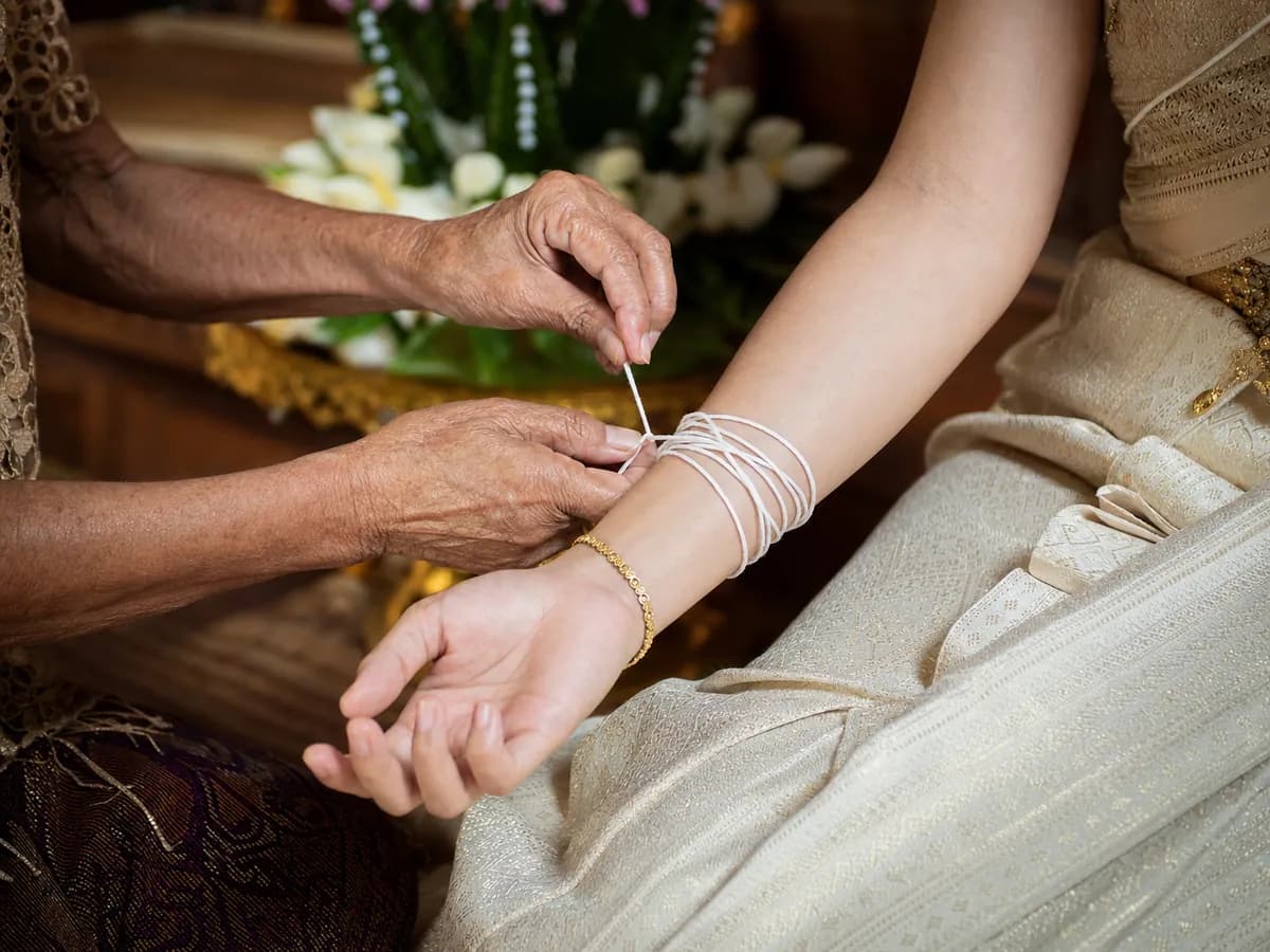 มือผู้ใหญ่ผูกด้ายฝ้ายสีขาวรอบข้อมือเจ้าสาวในพิธีบายศรีสู่ขวัญ