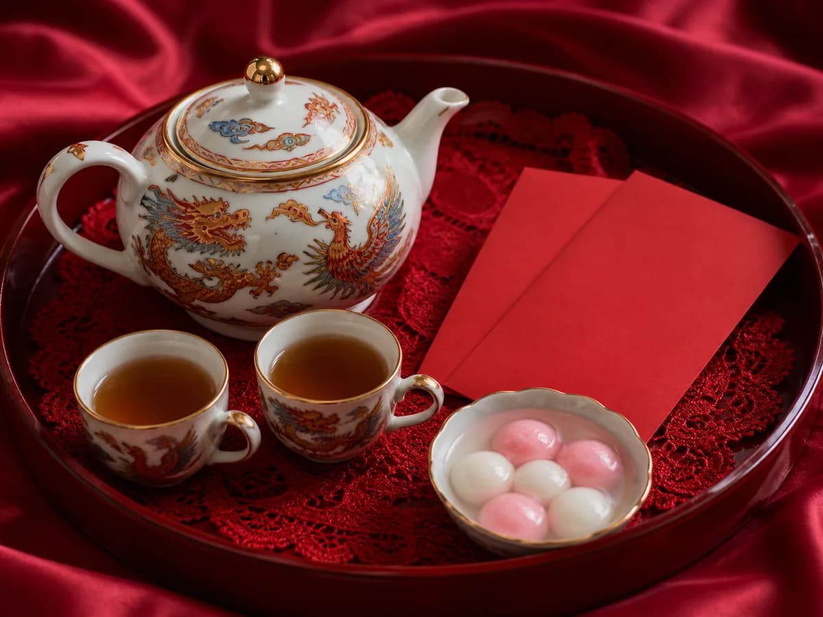 A dragon-and-phoenix tea set on a red lace-lined tray beside red envelopes and a bowl of khanom ee