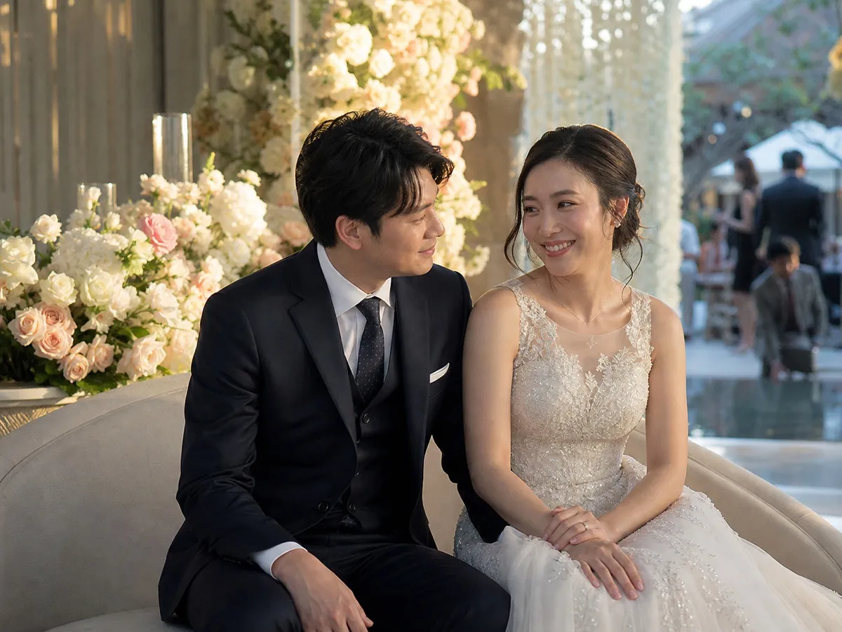 The couple relaxing on a sofa at their reception, smiling at each other while guests mingle behind